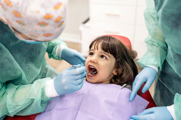 Young child in a dentist chair with a dentist looking in her mouth with a mirror and an assistance next to the child.